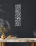 Mirror silver chrome Kalima calligraphy, Thuluth lines stacked in a tall vertical column, float-mounted clear of a stone-grey wall