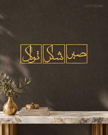 Set of three mirror gold acrylic panels reading Sabr, Shukr and Tawakkul in Arabic, each inside a slim rectangular border, hung in a row on a warm espresso wall