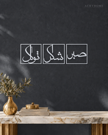 Set of three mirror silver acrylic panels reading Sabr, Shukr and Tawakkul in Arabic, each inside a slim rectangular border, hung in a row on a cool graphite wall