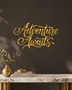 Adventure Awaits brush script wall art in mirror gold acrylic, float-mounted on a neutral wall with the flourishes catching warm light