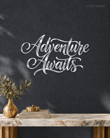 Adventure Awaits brush script wall art in mirror silver acrylic, float-mounted on a light wall above a desk