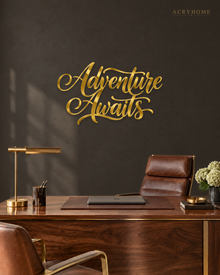 Adventure Awaits brush script wall art in mirror gold acrylic, float-mounted on a neutral wall with the flourishes catching warm light