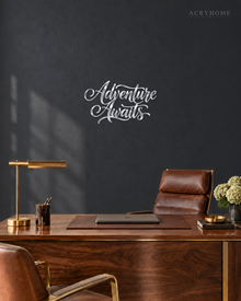 Adventure Awaits brush script wall art in mirror silver acrylic, float-mounted on a light wall above a desk