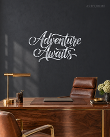 Adventure Awaits brush script wall art in mirror silver acrylic, float-mounted on a light wall above a desk