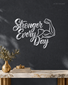 Stronger Every Day script wall art with flexed-arm icon in mirror silver acrylic, mounted above a dumbbell rack.