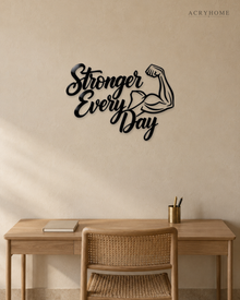 Stronger Every Day script wall art with flexed-arm icon in gloss piano black acrylic, float-mounted above a home gym bench.