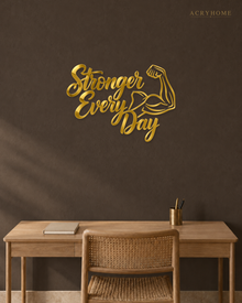 Stronger Every Day script wall art with flexed-arm icon in mirror gold acrylic, catching light on a pale gym wall.