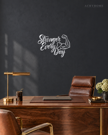 Stronger Every Day script wall art with flexed-arm icon in mirror silver acrylic, mounted above a dumbbell rack.
