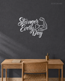 Stronger Every Day script wall art with flexed-arm icon in mirror silver acrylic, mounted above a dumbbell rack.
