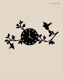 Gloss black acrylic wall clock with laser-cut branches, leaves and two small birds spreading asymmetrically either side of the dial, mounted on a pale wall.