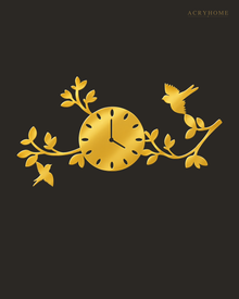 Mirror gold acrylic wall clock, branches, leaves and small birds cut around the clock face, catching warm light against a neutral wall.