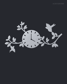 Mirror silver acrylic wall clock with a branch, leaf and bird design framing the dial, float-mounted on a light wall.