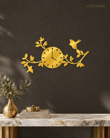 Mirror gold acrylic wall clock, branches, leaves and small birds cut around the clock face, catching warm light against a neutral wall.