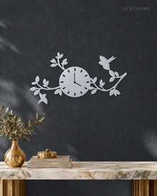 Mirror silver acrylic wall clock with a branch, leaf and bird design framing the dial, float-mounted on a light wall.