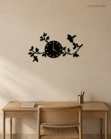 Gloss black acrylic wall clock with laser-cut branches, leaves and two small birds spreading asymmetrically either side of the dial, mounted on a pale wall.