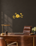 Mirror gold acrylic wall clock, branches, leaves and small birds cut around the clock face, catching warm light against a neutral wall.