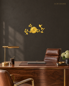 Mirror gold acrylic wall clock, branches, leaves and small birds cut around the clock face, catching warm light against a neutral wall.
