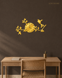 Mirror gold acrylic wall clock, branches, leaves and small birds cut around the clock face, catching warm light against a neutral wall.