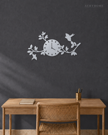 Mirror silver acrylic wall clock with a branch, leaf and bird design framing the dial, float-mounted on a light wall.