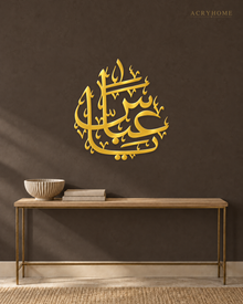 The same Arabic name calligraphy piece in mirror gold acrylic, catching warm light on a light-toned wall.