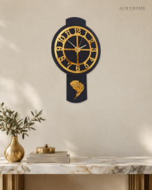 Pendulum wall clock in black and gold mirror acrylic, round dial above a long drop, on a pale wall.